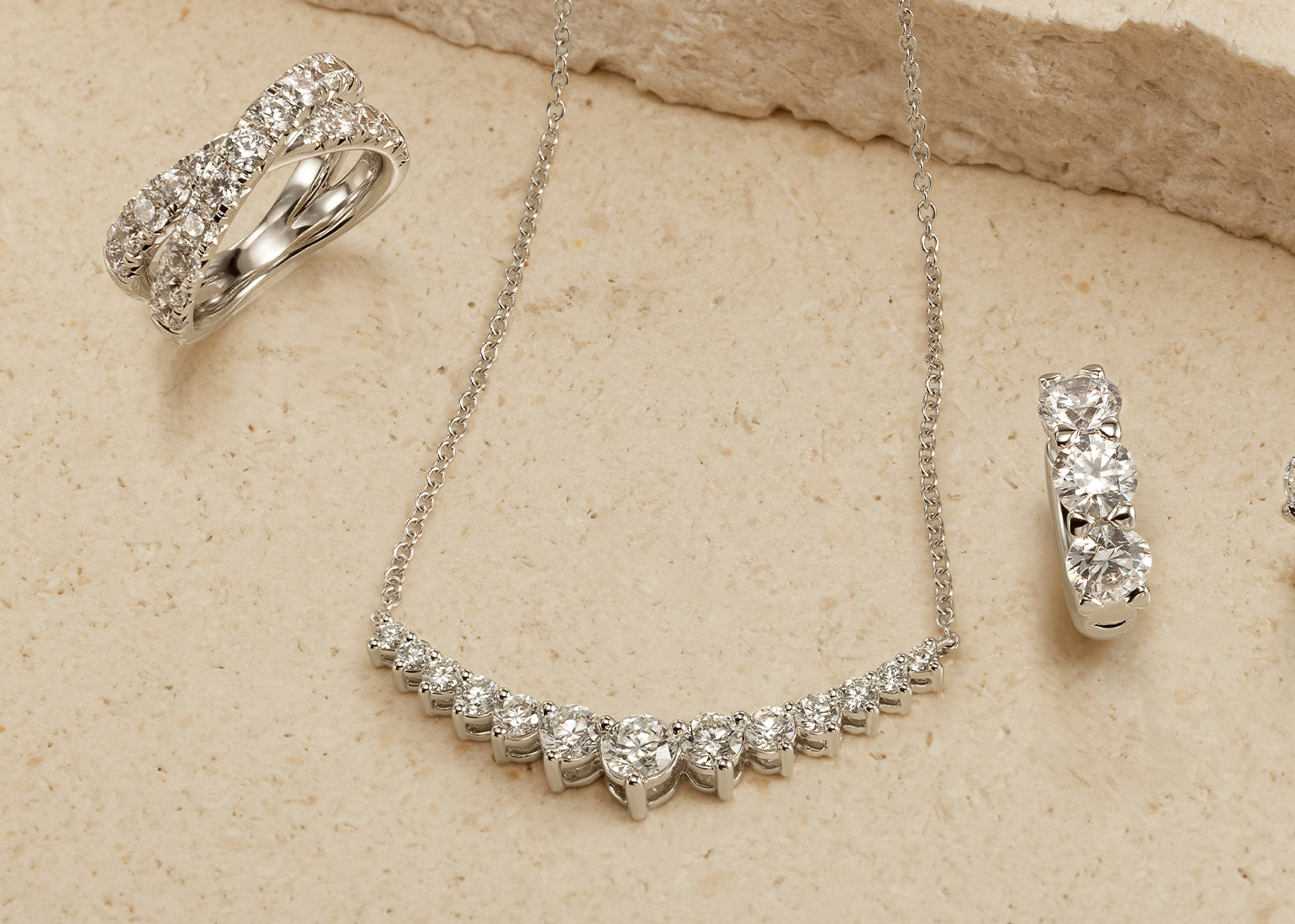 An landscape image featuring a diamond necklace and three diamond rings with a neutral background